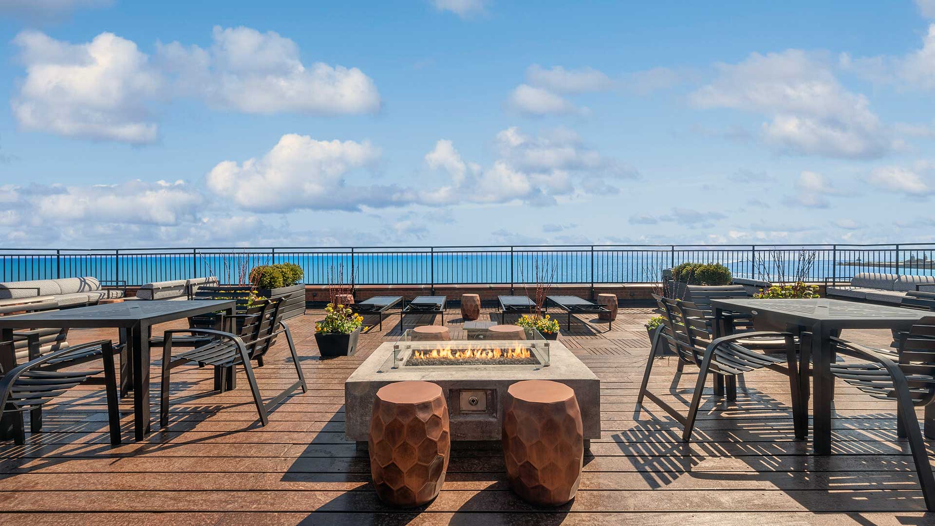 Looking across the rooftop deck firepit at Lake Michigan on a partly cloudy day. Outdoor dining sets sit on either side.