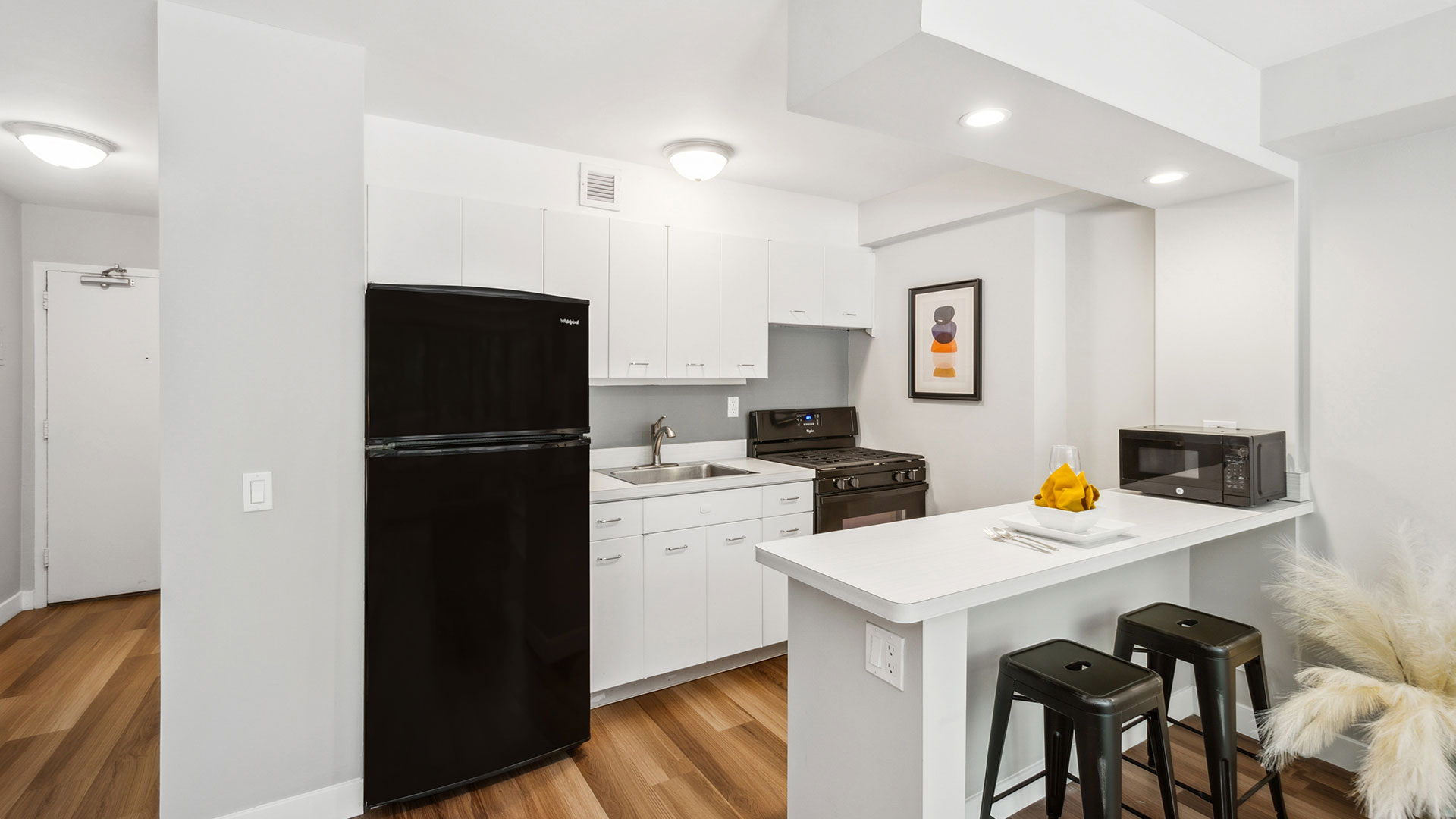 Modern kitchen with white cabinets, black appliances, a breakfast bar with two stools, and a wooden floor. A microwave and a framed art piece are visible, enhancing the minimalist decor.